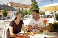 Bild: Ein Mann und eine Frau, die auf dem Marktplatz einen Brotzeitteller essen und Bier trinken. Im Hintergrund ist die Kirche zu sehen.